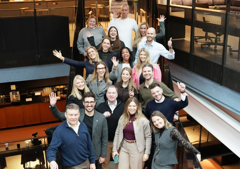 Brandpie team members gathered together in a modern office, smiling and waving at the camera from multiple levels of a staircase.