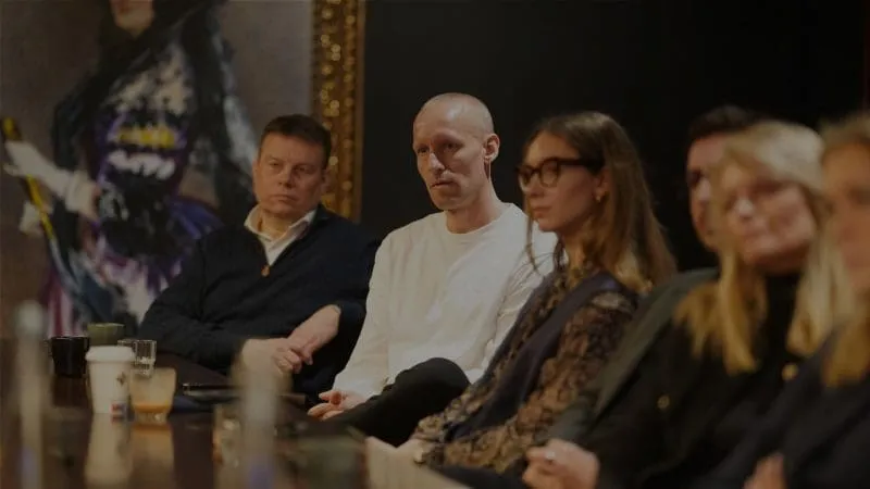 A group of people sit around a table in a meeting or discussion, listening attentively. A man in a white shirt is in central focus, with other attendees seated beside him and drinks on the table.