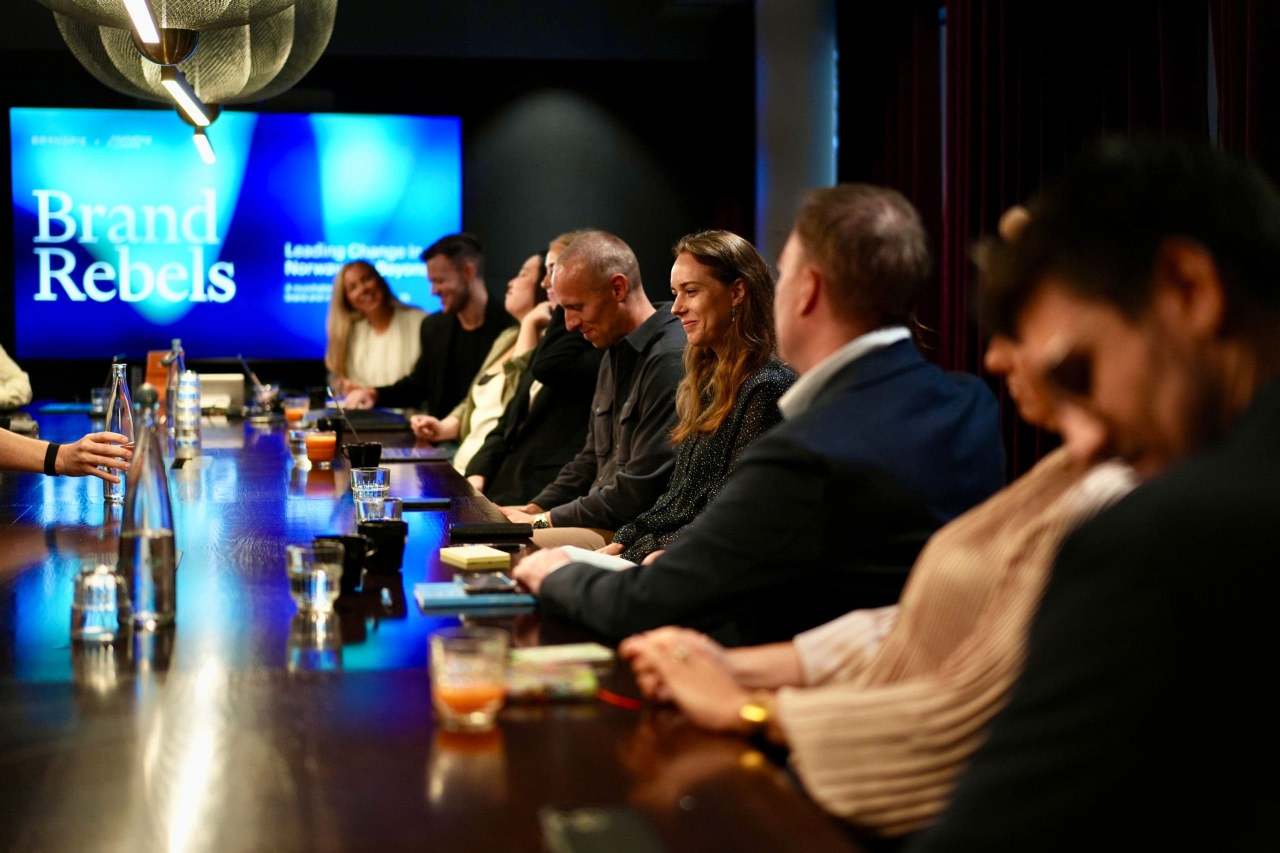 People seated around a conference table at a Brand Rebels event, with a large “Brand Rebels” presentation screen visible in the background.