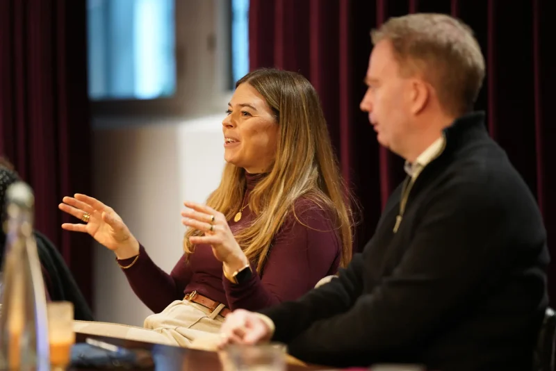A woman gestures with both hands while speaking during a seated panel discussion, with a man beside her listening.