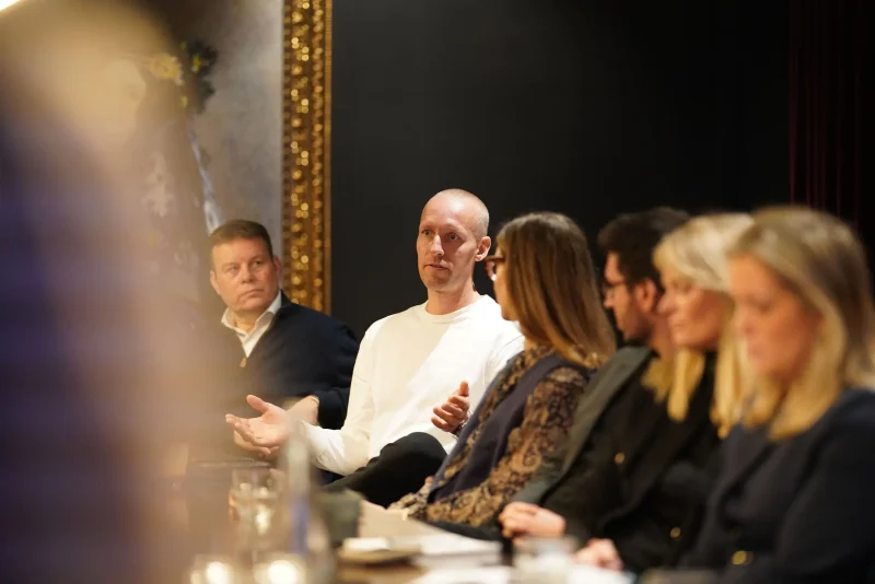 A bald man in a white shirt gestures while speaking during a roundtable discussion, as several other participants seated beside him listen.