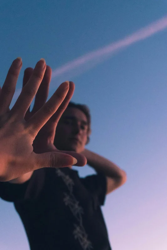 Person reaching an open hand toward the camera against a blue and pink sky, with their face softly out of focus in the background.